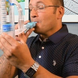 A person wearing glasses and a dotted polo shirt uses a handheld lung capacity tester, blowing into the mouthpiece while holding the device upright. A framed leaf artwork is visible on the wall in the background.