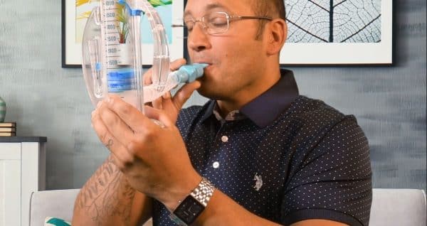 A person wearing glasses and a dotted polo shirt uses a handheld lung capacity tester, blowing into the mouthpiece while holding the device upright. A framed leaf artwork is visible on the wall in the background.